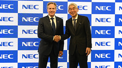 US Foreign Secretary Blinken poses for a photo with NEC President and CEO Takayuki Morita in front of a wall of blue and white NEC logos to promote Open RAN